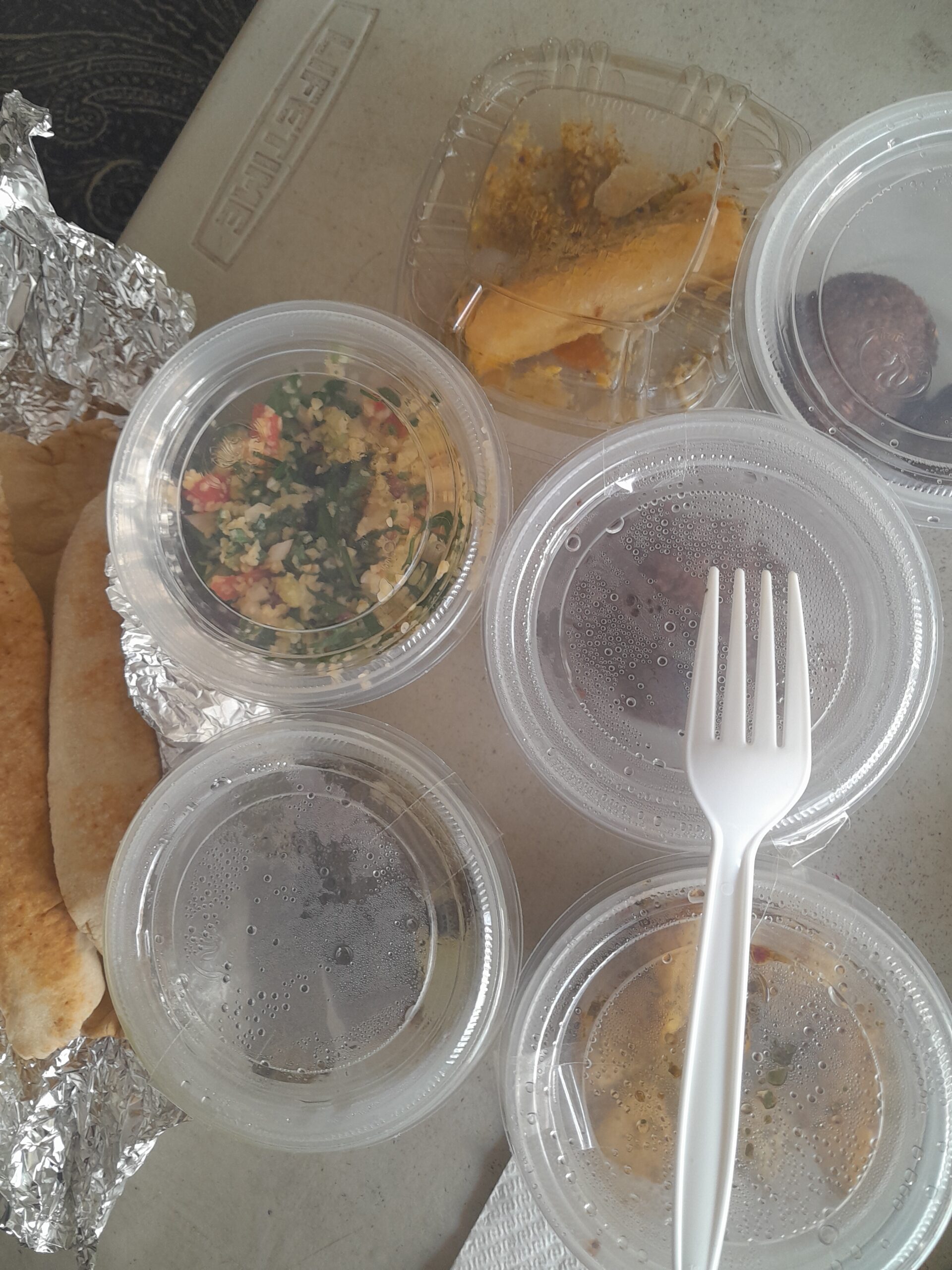 Comida para llevar que incluye comida libanesa en varios recipientes de plástico, pan pita en papel aluminio y un tenedor de plástico blanco sobre una superficie de color claro, perfecto para disfrutar de los sabores de León Gto. Takeout meal featuring comida libanesa in several plastic containers, pita bread in foil, and a white plastic fork on a light-colored surface, perfect for enjoying flavors from León Gto.